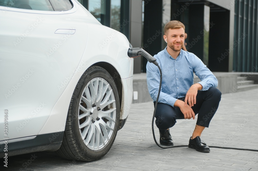 Hansome bearded guy sitting near his new modern electric car and holding plug of the charger, while car is charging at the charging station.