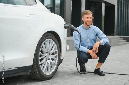 Hansome bearded guy sitting near his new modern electric car and holding plug of the charger, while car is charging at the charging station. © Serhii