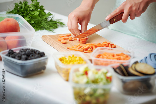 Hands of young female chopping fresh carrot on wooden board while preparing vegetables for freezing
