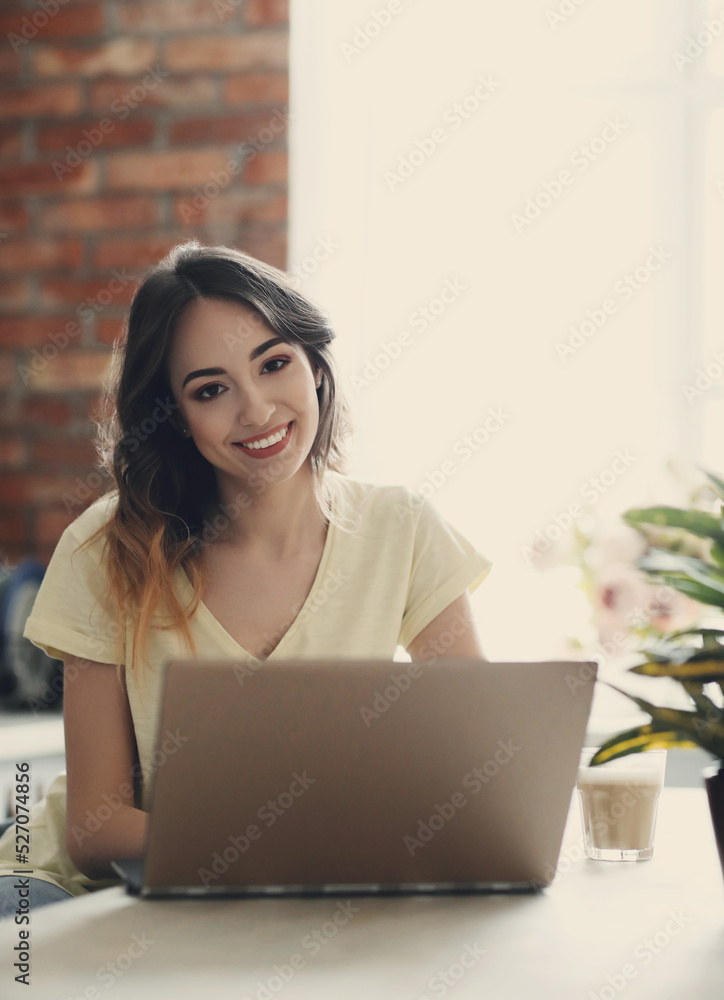 Young, charming, brunette girl sitting in the kitchen with a computer and looking into the camera Young, charming, brunette girl sitting in the kitchen with a computer and looking into the camera
