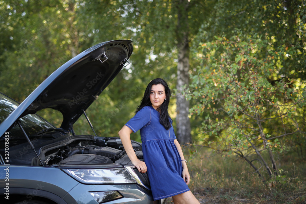 A young brunette woman sitting on the hood of her broken car and waiting help