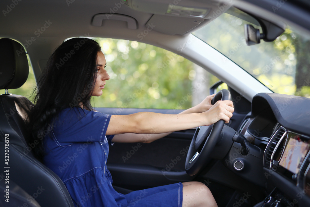 Photo of a young, brunette woman in a blue dress sitting behind the wheel of her car 