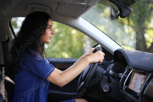 Photo of a young, brunette woman in a blue dress sitting behind the wheel of her car 