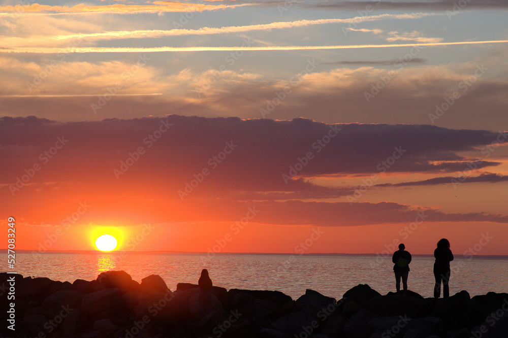 Sunset at Cape Cod beach. Dark outlines (silhouettes) of unrecognizable people on breakwater rocks viewing colorful sky and yellow sun just above horizon with reflection on water.