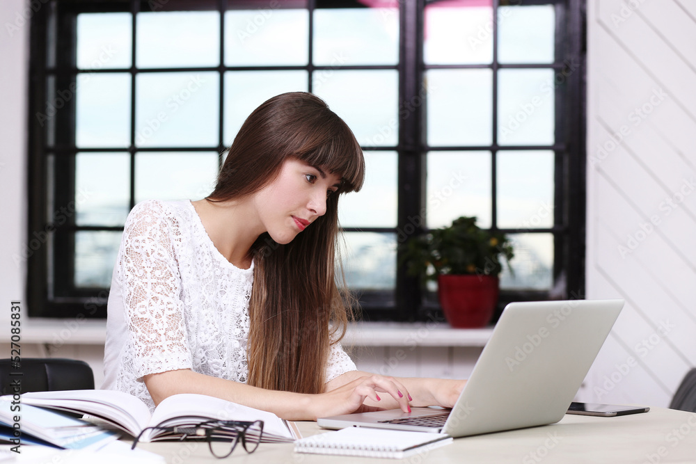 Beautiful, sweet, young girl with long hair sitting and working on the computer against windowsill. Beautiful, sweet, young girl with long hair sitting and working on the computer against windowsill.