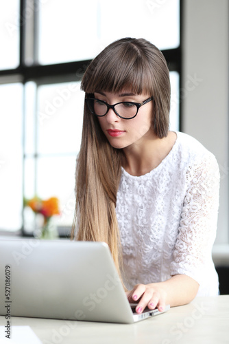 Young, beautiful, cute girl with bangs in glasses studying on the computer against windowsill.