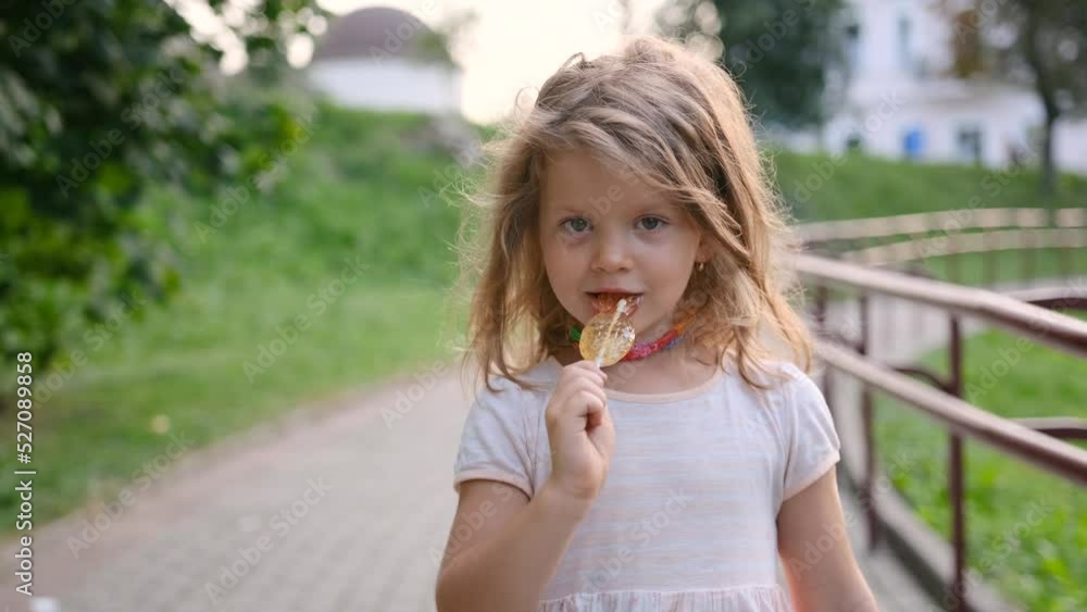 A little girl is walking down the street eating candy on a stick close-up. A child with sweets. Happy child. A little girl is walking down the street eating candy on a stick close-up. A child with sweets. Happy child.