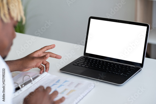Online conference. Office man. Digital mockup. Unrecognizable black guy talking to laptop blank screen checking documents sitting desk in light room interior.