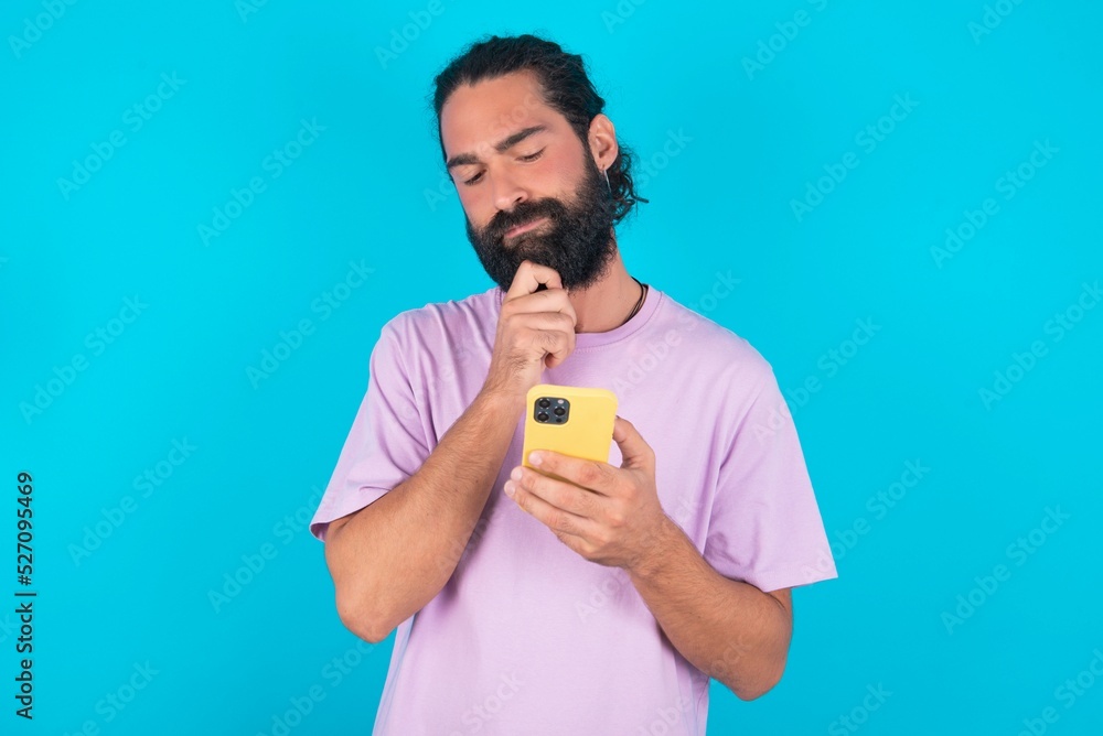 Thoughtful happy young bearded man wearing violet T-shirt over blue studio background hold look phone, copyspace Thoughtful happy young bearded man wearing violet T-shirt over blue studio background hold look phone, copyspace