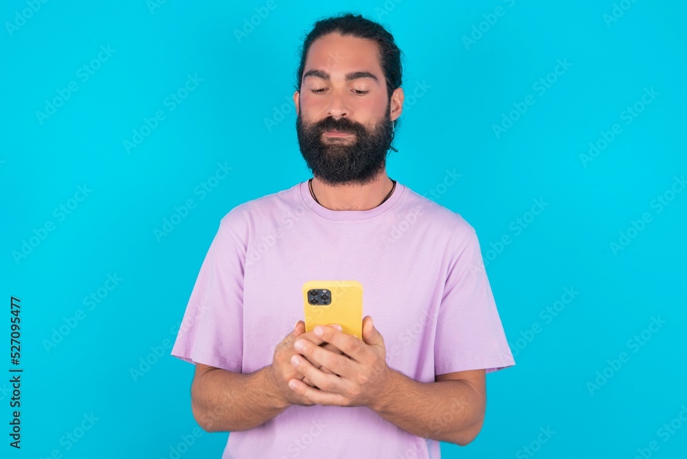 Portrait of serious confident young bearded man wearing violet T-shirt over blue studio background holding phone in two hands Portrait of serious confident young bearded man wearing violet T-shirt over blue studio background holding phone in two hands
