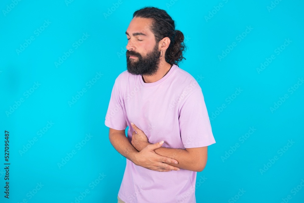young bearded man wearing violet T-shirt over blue studio background got stomachache young bearded man wearing violet T-shirt over blue studio background got stomachache