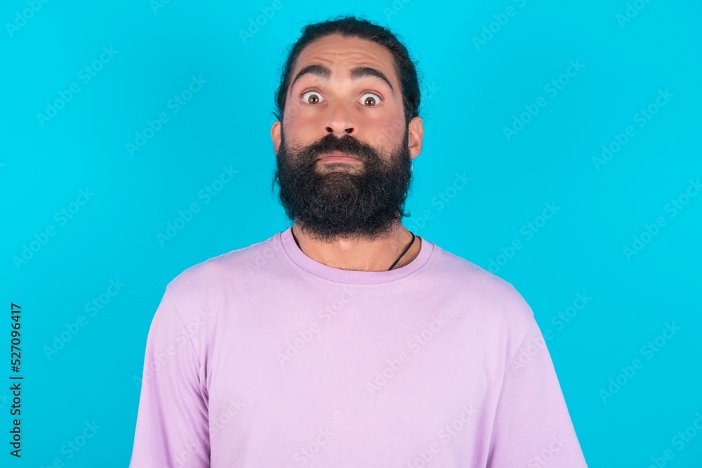 Stunned young bearded man wearing violet T-shirt over blue studio background stares reacts on shocking news. Astonished latin man holds breath Stunned young bearded man wearing violet T-shirt over blue studio background stares reacts on shocking news. Astonished latin man holds breath