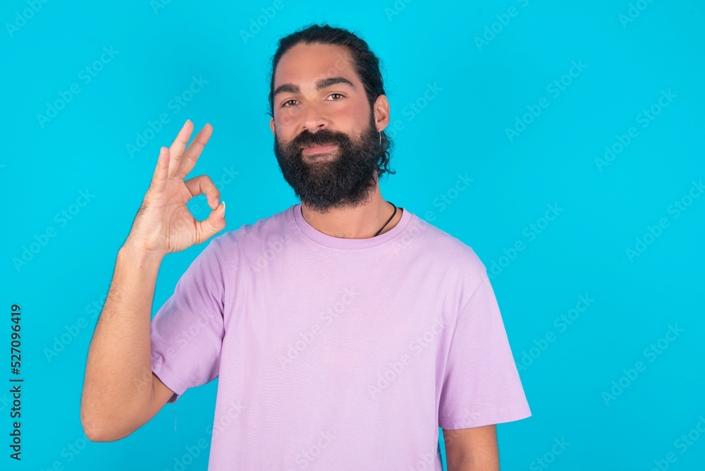 young bearded man wearing violet T-shirt over blue studio background hold hand arm okey symbol toothy approve advising novelty news young bearded man wearing violet T-shirt over blue studio background hold hand arm okey symbol toothy approve advising novelty news