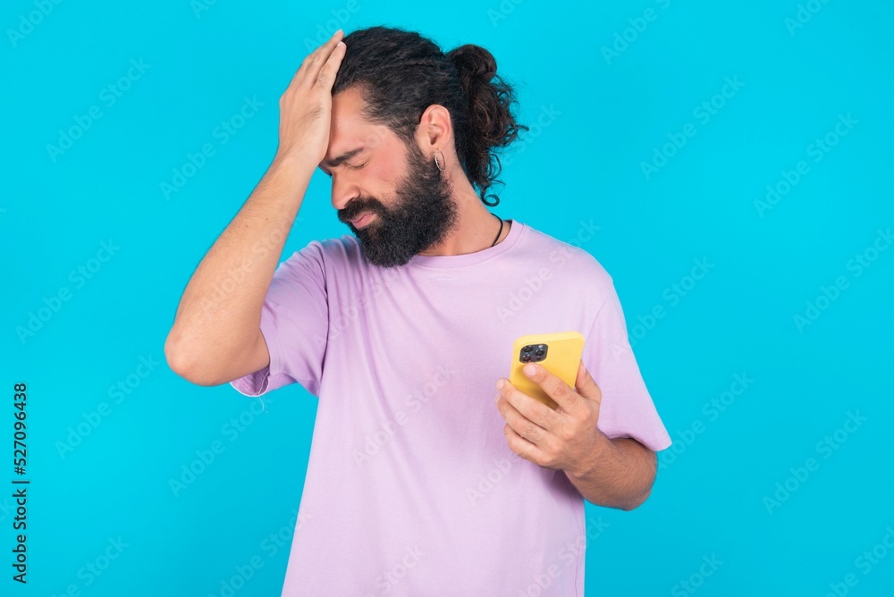 Upset depressed young bearded man wearing violet T-shirt over blue studio background makes face palm as forgot about something important holds mobile phone expresses sorrow and regret blames Upset depressed young bearded man wearing violet T-shirt over blue studio background makes face palm as forgot about something important holds mobile phone expresses sorrow and regret blames