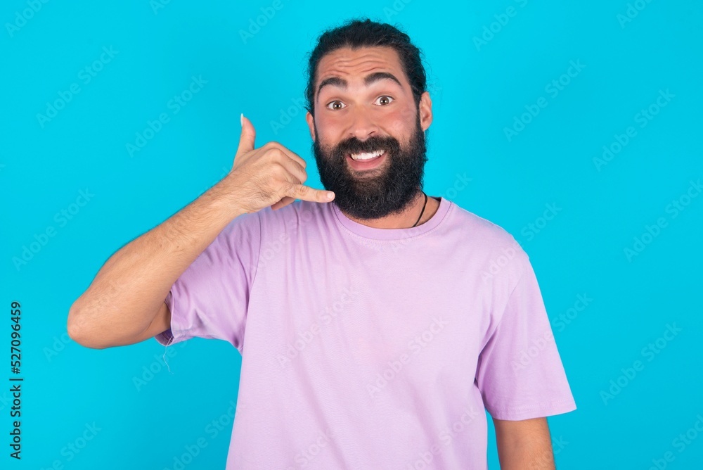 young bearded man wearing violet T-shirt over blue studio background makes phone gesture, says call me back again, has glad expression. young bearded man wearing violet T-shirt over blue studio background makes phone gesture, says call me back again, has glad expression.