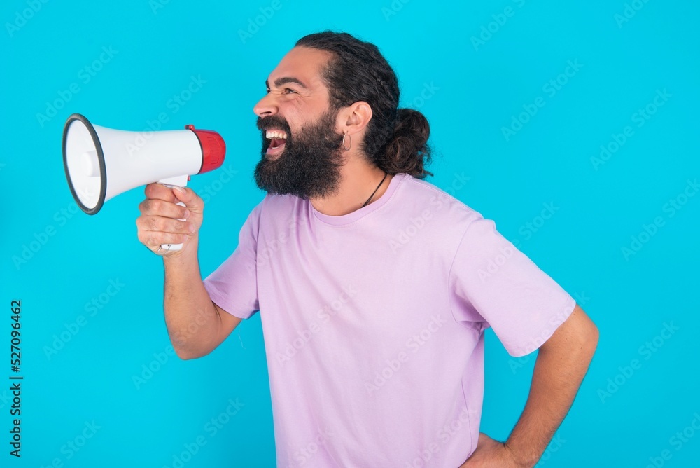 Funny young bearded man wearing violet T-shirt over blue studio background People sincere emotions lifestyle concept. Mock up copy space. Screaming in megaphone. Funny young bearded man wearing violet T-shirt over blue studio background People sincere emotions lifestyle concept. Mock up copy space. Screaming in megaphone.