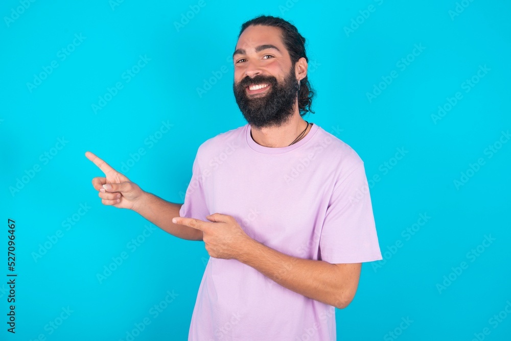 young bearded man wearing violet T-shirt over blue studio background points at copy space indicates for advertising gives right direction young bearded man wearing violet T-shirt over blue studio background points at copy space indicates for advertising gives right direction