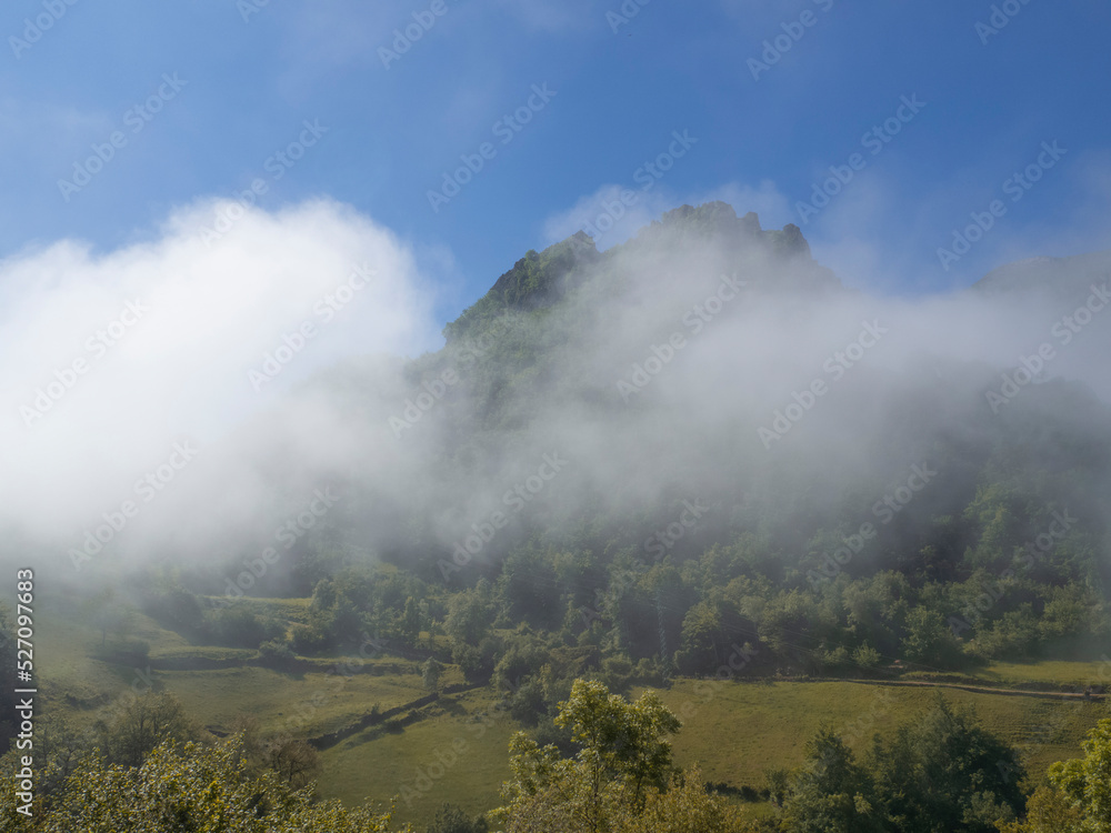 Paisaje de montañas con niebla blanca, baja visibilidad, con árboles sobre fondo del cielo azul en verano de 2021 en Asturias.