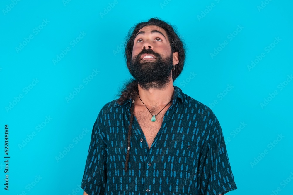 young bearded man wearing blue shirt over blue studio background looking up as he sees something strange. young bearded man wearing blue shirt over blue studio background looking up as he sees something strange.