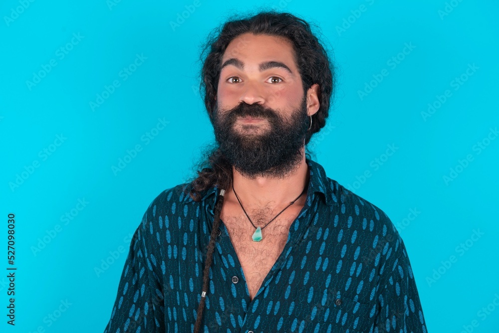 young bearded man wearing blue shirt over blue studio background puffing cheeks with funny face. Mouth inflated with air, crazy expression. young bearded man wearing blue shirt over blue studio background puffing cheeks with funny face. Mouth inflated with air, crazy expression.