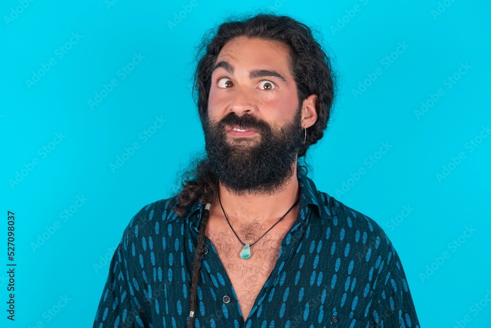 young bearded man wearing blue shirt over blue studio background crosses eyes, puts lips, makes grimace with awkward expression has fun alone, plays fool. young bearded man wearing blue shirt over blue studio background crosses eyes, puts lips, makes grimace with awkward expression has fun alone, plays fool.
