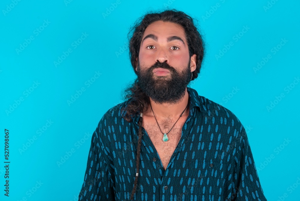 young bearded man wearing blue shirt over blue studio background, keeps lips as going to kiss someone, has glad expression, grimace face. Standing indoors. Beauty concept. young bearded man wearing blue shirt over blue studio background, keeps lips as going to kiss someone, has glad expression, grimace face. Standing indoors. Beauty concept.