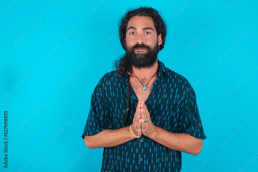 young bearded man wearing blue shirt over blue studio background keeps palms pressed together in front of her having regretful look, asking for forgiveness. Forgive me please.