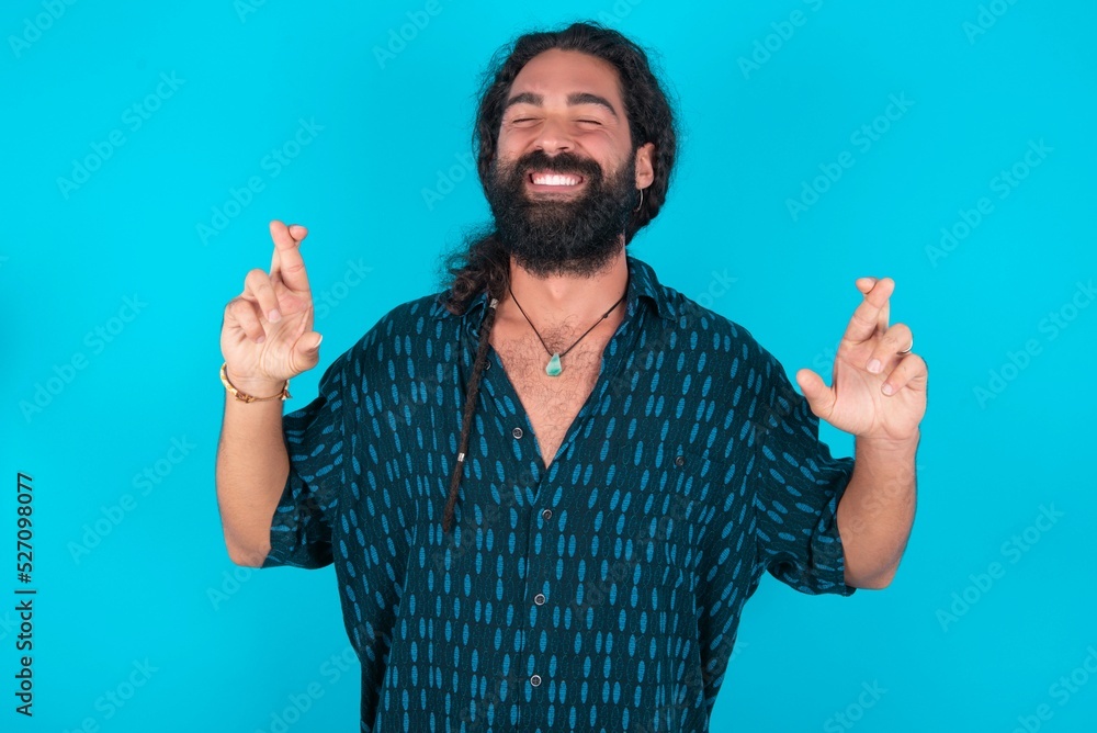 Joyful young bearded man wearing blue shirt over blue studio background clenches teeth, raises fingers crossed, makes desirable wish, waits for good news, I have to win. Joyful young bearded man wearing blue shirt over blue studio background clenches teeth, raises fingers crossed, makes desirable wish, waits for good news, I have to win.