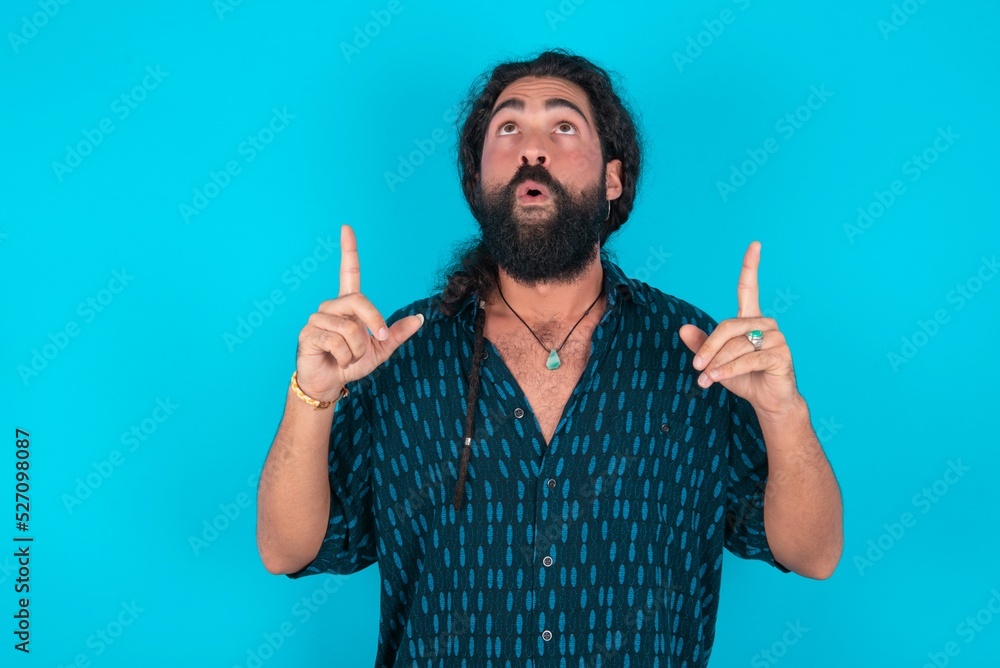 young bearded man wearing blue shirt over blue studio background being amazed and surprised looking and pointing up with fingers showing something strange. young bearded man wearing blue shirt over blue studio background being amazed and surprised looking and pointing up with fingers showing something strange.