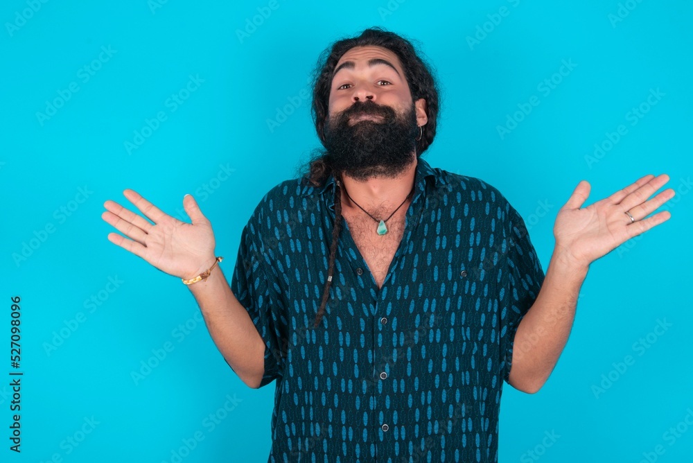 Maybe yes or no. Doubtful young bearded man wearing blue shirt over blue studio background shrugs shoulders in bewilderment, tries to make decision puzzled what he wants. Hesitation and uncertainty. Maybe yes or no. Doubtful young bearded man wearing blue shirt over blue studio background shrugs shoulders in bewilderment, tries to make decision puzzled what he wants. Hesitation and uncertainty.