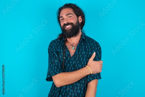 People, lifestyle, youth and happiness concept. Shy pretty young bearded man wearing blue shirt over blue studio background, feeling happy hugging herself.