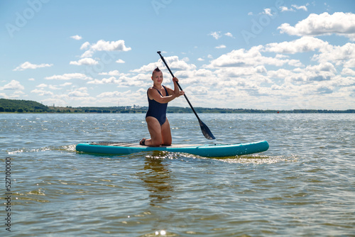 a woman in a closed swimsuit with kneeling on a SUP board with a paddle floats on the water against the blue sky.