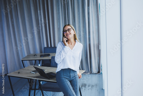 A successful smiling business woman is standing in the office by the window with a phone in her hands. A female manager in glasses and a white shirt is talking on the phone