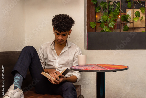 Latin man with curly hair, in a room, sitting on a sofa reading a book and a cup next to a table with a cup of coffee