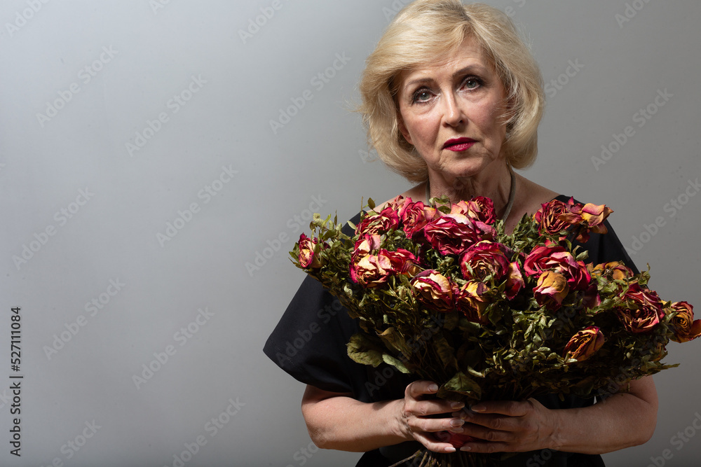 Sad elderly woman touching bouquet of withered rose flowers. Gray haired mature lady in depression. Concept of old aging. Studio shoot isolated on gray background. Copy space Sad elderly woman touching bouquet of withered rose flowers. Gray haired mature lady in depression. Concept of old aging. Studio shoot isolated on gray background. Copy space