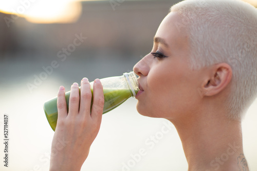 Portrait of young smiling millenial european short haired woman with green smoothie. Beautiful happy blonde girl outdoor.