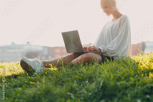 Portrait of young smiling millenial european short haired woman using laptop at green grass meadow in park. Beautiful happy blonde girl outdoor. Summer fashion female clothing.