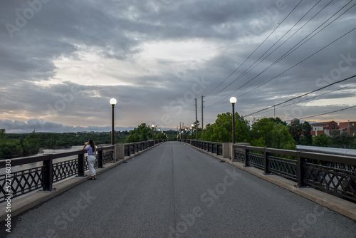 View down bridge roadway deck at dusk with lone woman looking at the river below, lit street lamps on bridge railing, dramatic cloudy sky