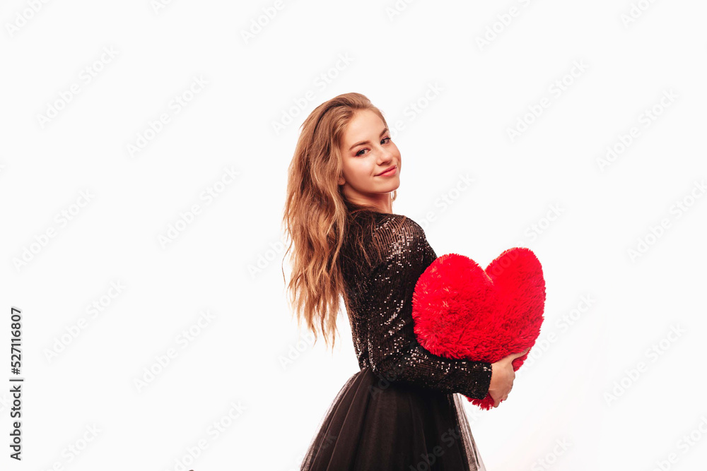 Smiling teenage girl with blond curly hair and in a black dress holding a large plush heart, isolated on a white background. Copy space. Smiling teenage girl with blond curly hair and in a black dress holding a large plush heart, isolated on a white background. Copy space.