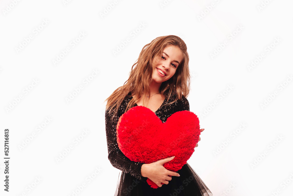 Smiling teenage girl with blond curly hair and in a black dress holding a large plush heart, isolated on a white background. Copy space. Smiling teenage girl with blond curly hair and in a black dress holding a large plush heart, isolated on a white background. Copy space.