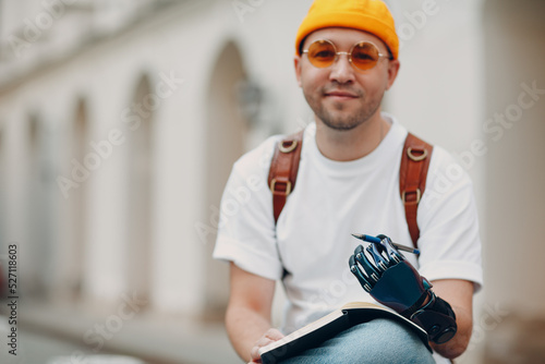 Young disabled man writes in notebook with pen in artificial prosthetic hand at city street outdoors