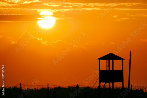 Fence with barbed wire and an old watchtower background bright sun and scarlet sunset.