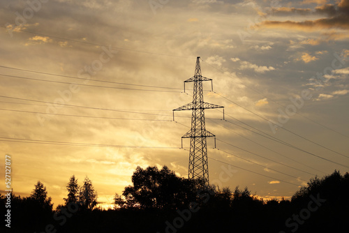 Silhouette of high voltage tower with electrical wires on background of sunset sky and dark clouds. Electricity transmission lines in evening forest, power supply concept
