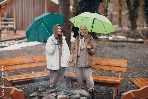 Women relaxing at bonfire with umbrella at glamping camping. Modern vacation lifestyle concept
