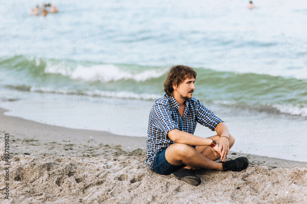 A handsome, young hippie man in a shirt and shorts sits on the sand against the background of the blue sea, ocean, relaxing, meditating on the beach.