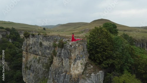 A girl in a red dress stands on the edge of a cliff, summer, mountains and forest, a seed from a drone