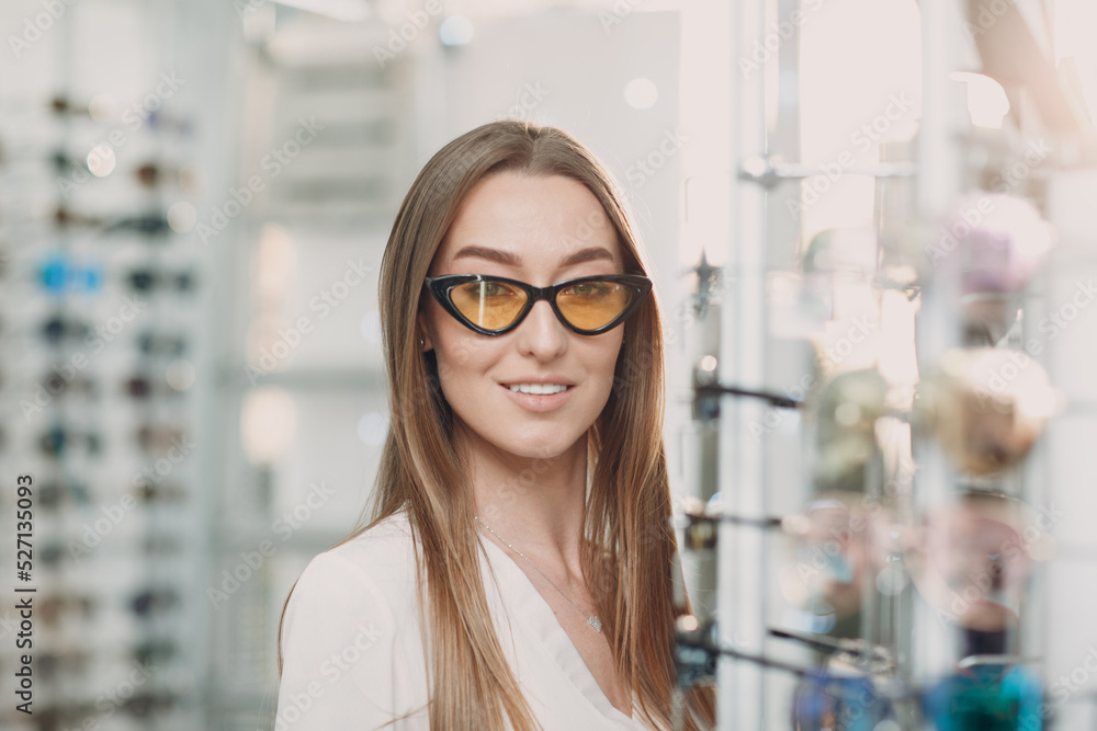 Close up of gorgeous young woman smiling picking and choosing glasses at the optician corner at the shopping mall. Happy beautiful woman buying eyewear eyeglasses at the optometrist.