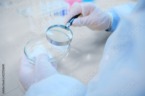 Selective focus: Loupe with magnifying glass in a scientist pharmacologist hands, examining pharmacological substance on Petri dish, sitting at table with empty test tubes on tripod in medical biolab.