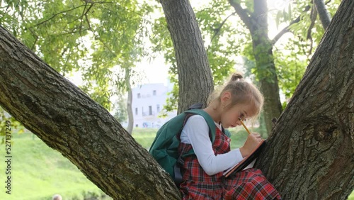 a schoolgirl girl in a red dress draws in a notebook with a pencil sitting on the branches of a tree.
