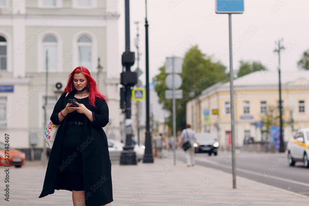 European plus size woman walking outdoor street city with mobile cellular phone. Young red pink haired body positive girl using cellphone smartphone European plus size woman walking outdoor street city with mobile cellular phone. Young red pink haired body positive girl using cellphone smartphone